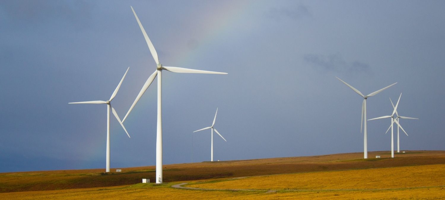 5 wind turbines sitting on a hill in the sunshine with a rainbow in the sky