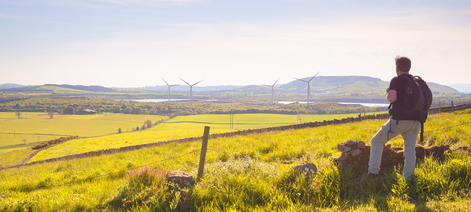 A man stands with a rucksack looking out over a field and in the distance is Kinglassie Wind Farm which has funding administered by Foundation Scotland