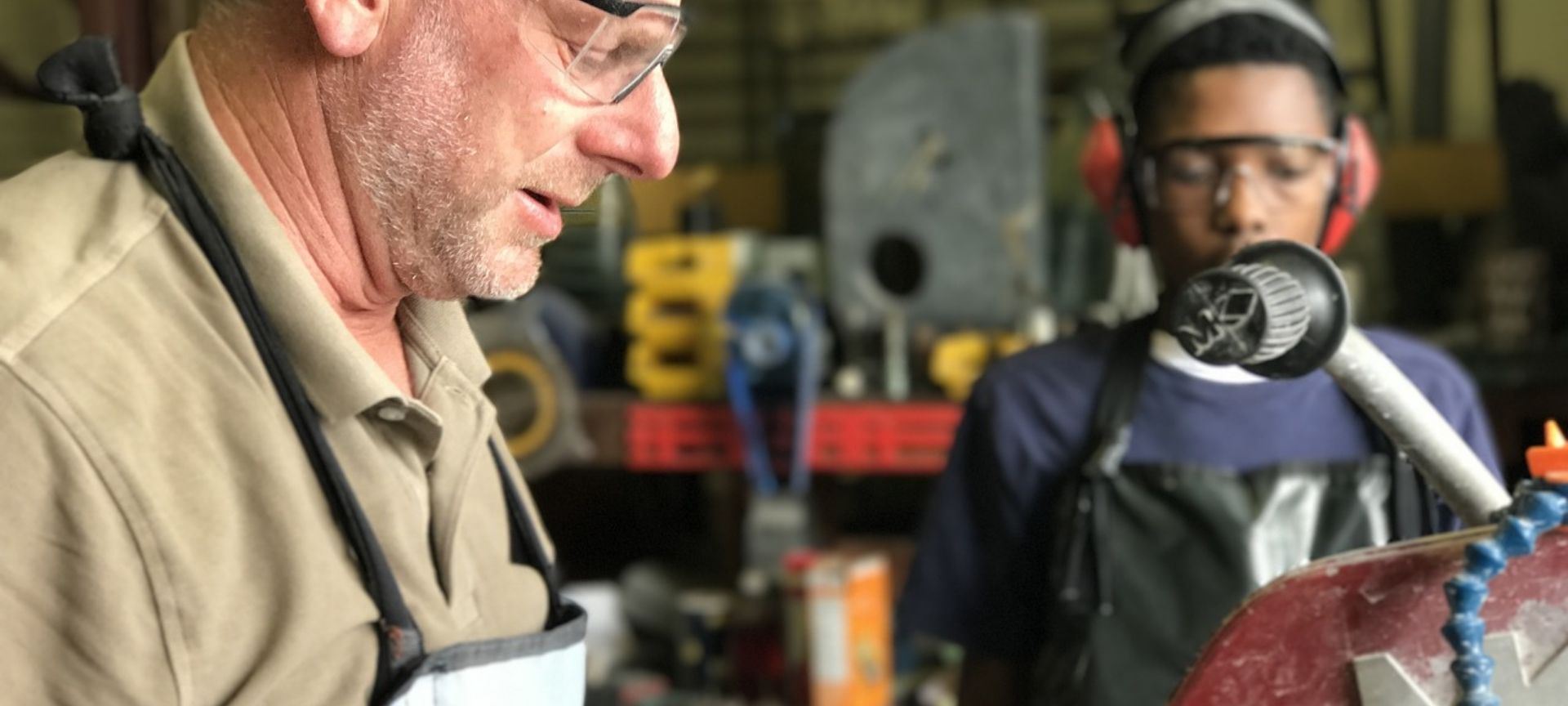 A photo of two men, the older man is working with machinery and the other watching.